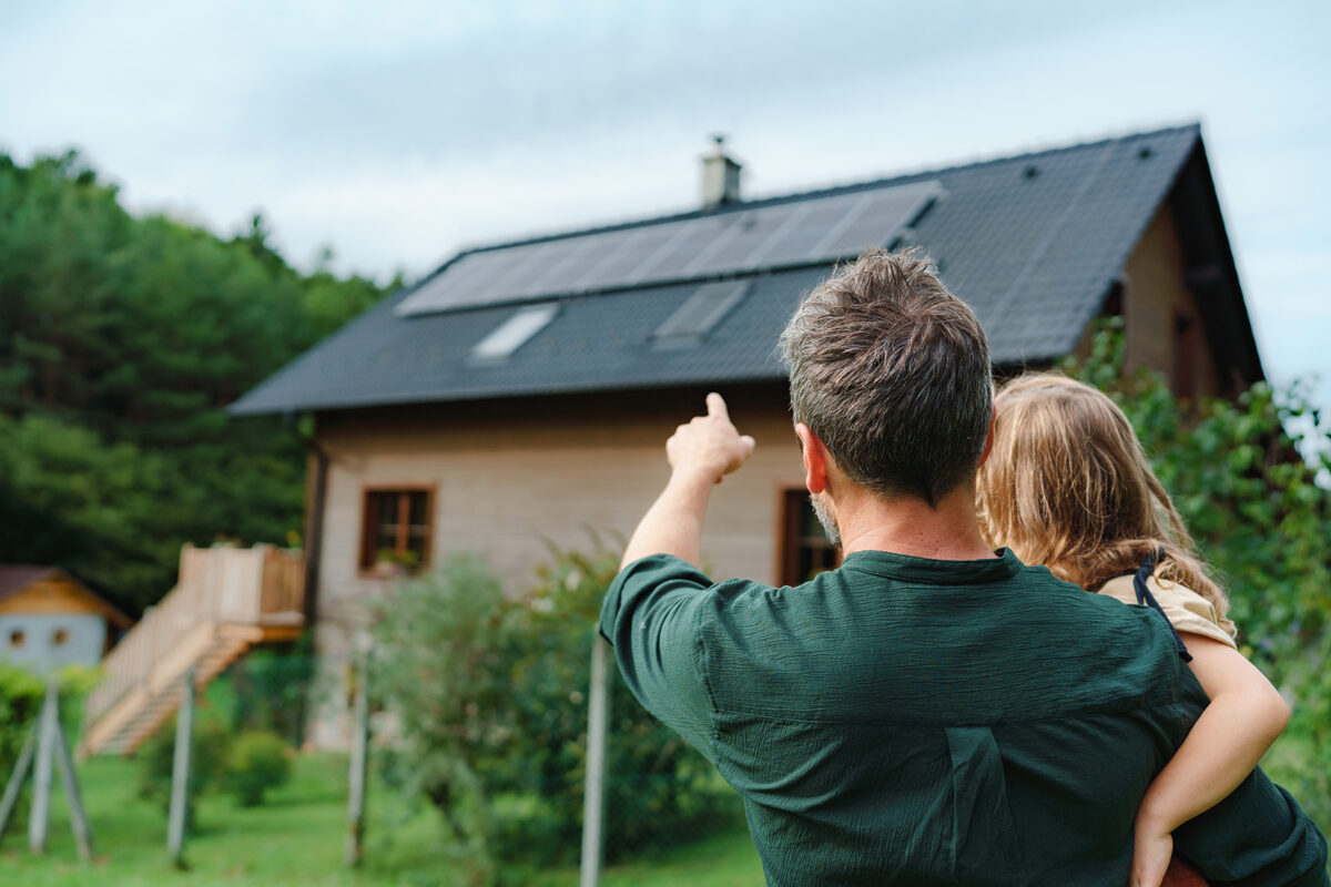 Ein Vater mit Kind auf dem Arm erklärt seiner Tocher die Photovoltaikanlage welche auf dem Hausdach installiert ist, auf welches er mit dem Finger zeigt.
